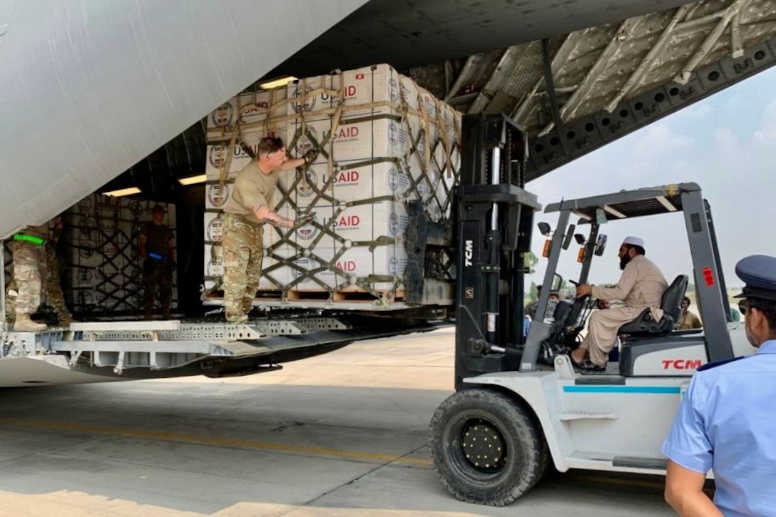 A man driving a forklift moves pallets of supplies and unloads them from a large plane while a soldier on board gives directions.