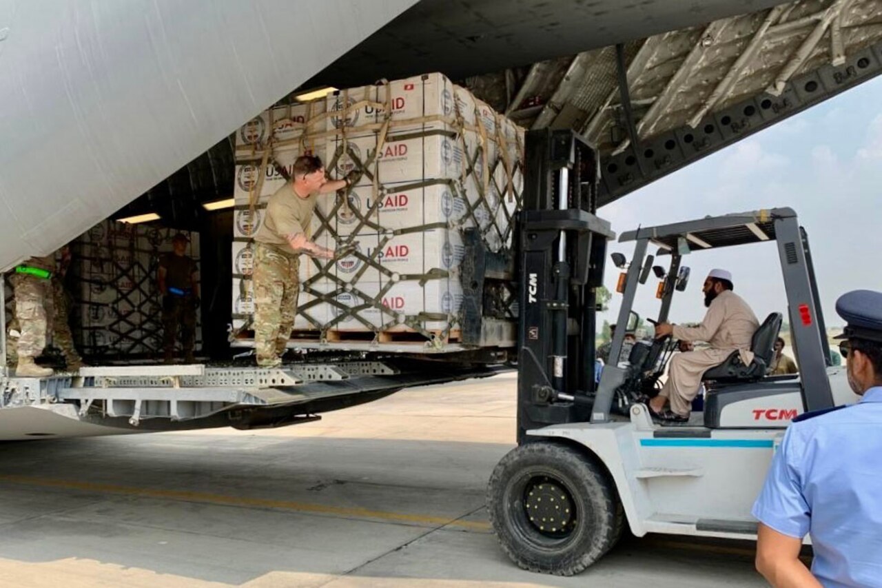 A man driving a forklift moves pallets of supplies and unloads them from a large plane while a soldier on board gives directions.