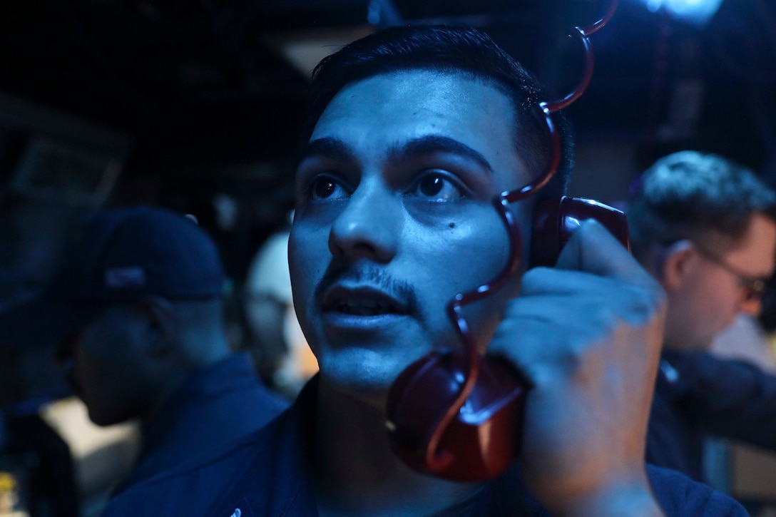 A sailor holds a red phone while looking upwards in a blue-lit room.