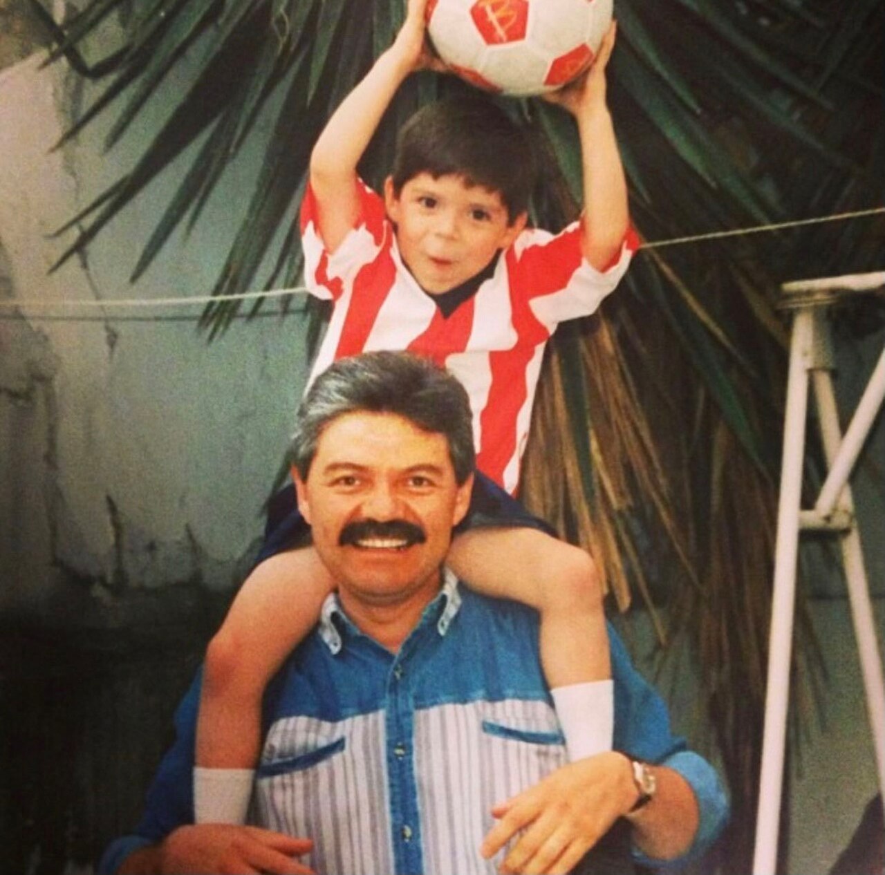 A young boy holding a soccer ball sits on his father's shoulders.