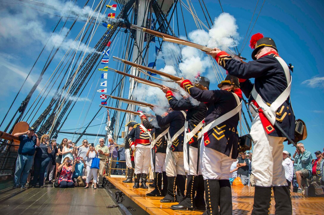 Personnel dressed as Marines in 1812 fire weapons aboard a ship as people watch.