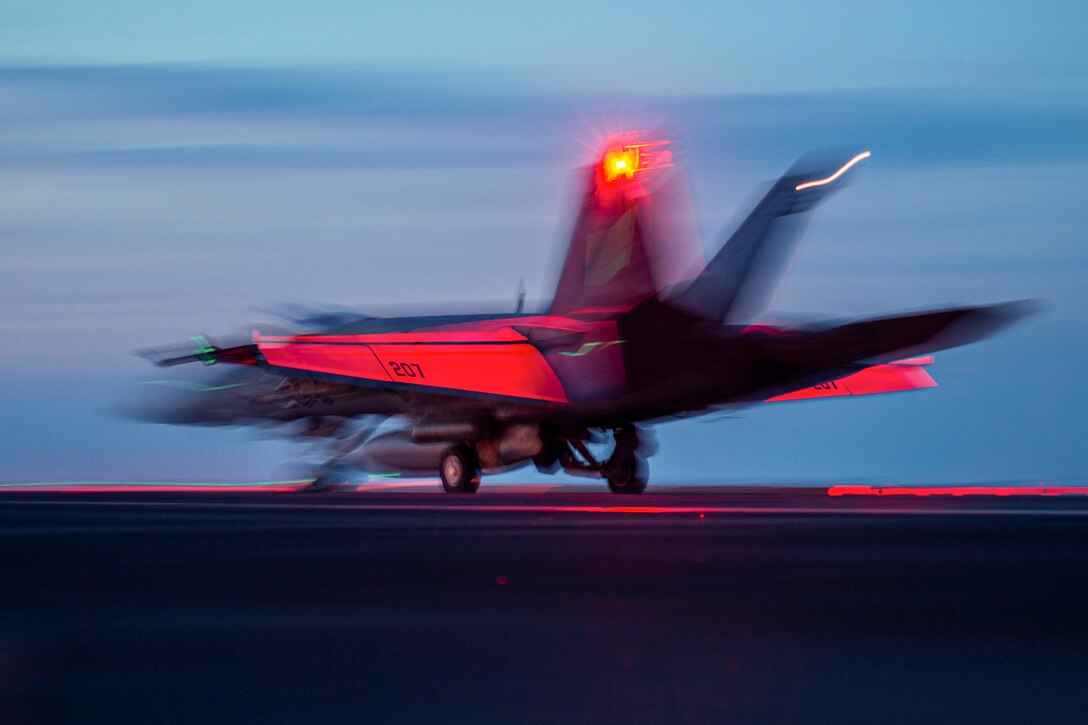 An aircraft launches from a ship illuminated by red light.