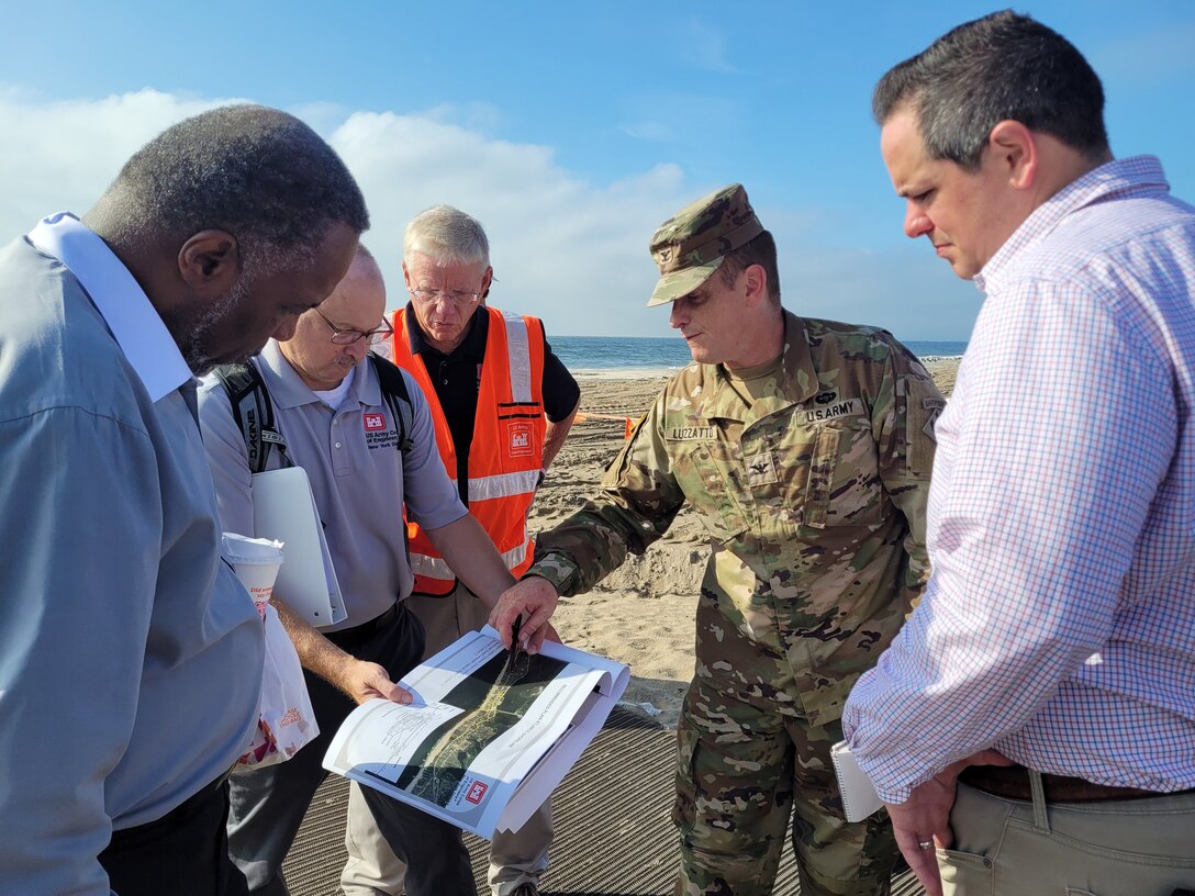 Five men stand on Rockaway Beach reviewing plans held by one of the men for U.S. Army Corps of Engineers projects. The ocean appears in the photo's background.