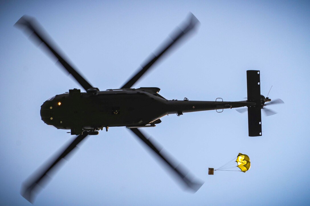 A look underneath an airborne helicopter as a package descends attached to a parachute below.