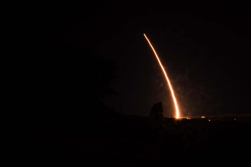 The flames from a rocket illuminate its smoke trail against the night sky.