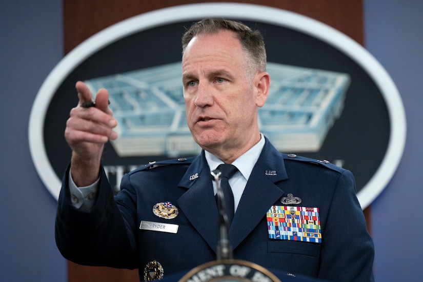 A man in a military uniform, standing behind a lectern, points to a person in the unseen audience.