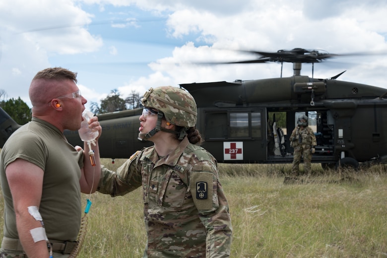 A soldier grabs hold of another soldier, both yelling at each other, one with an IV bag in his hand with a running medical helicopter in the background.