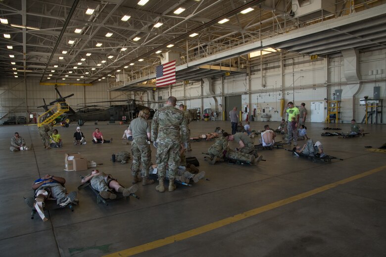 Uniformed military personnel stand in a hangar with patients in litters with a UH-60 Blackhawk helicopter in the background.