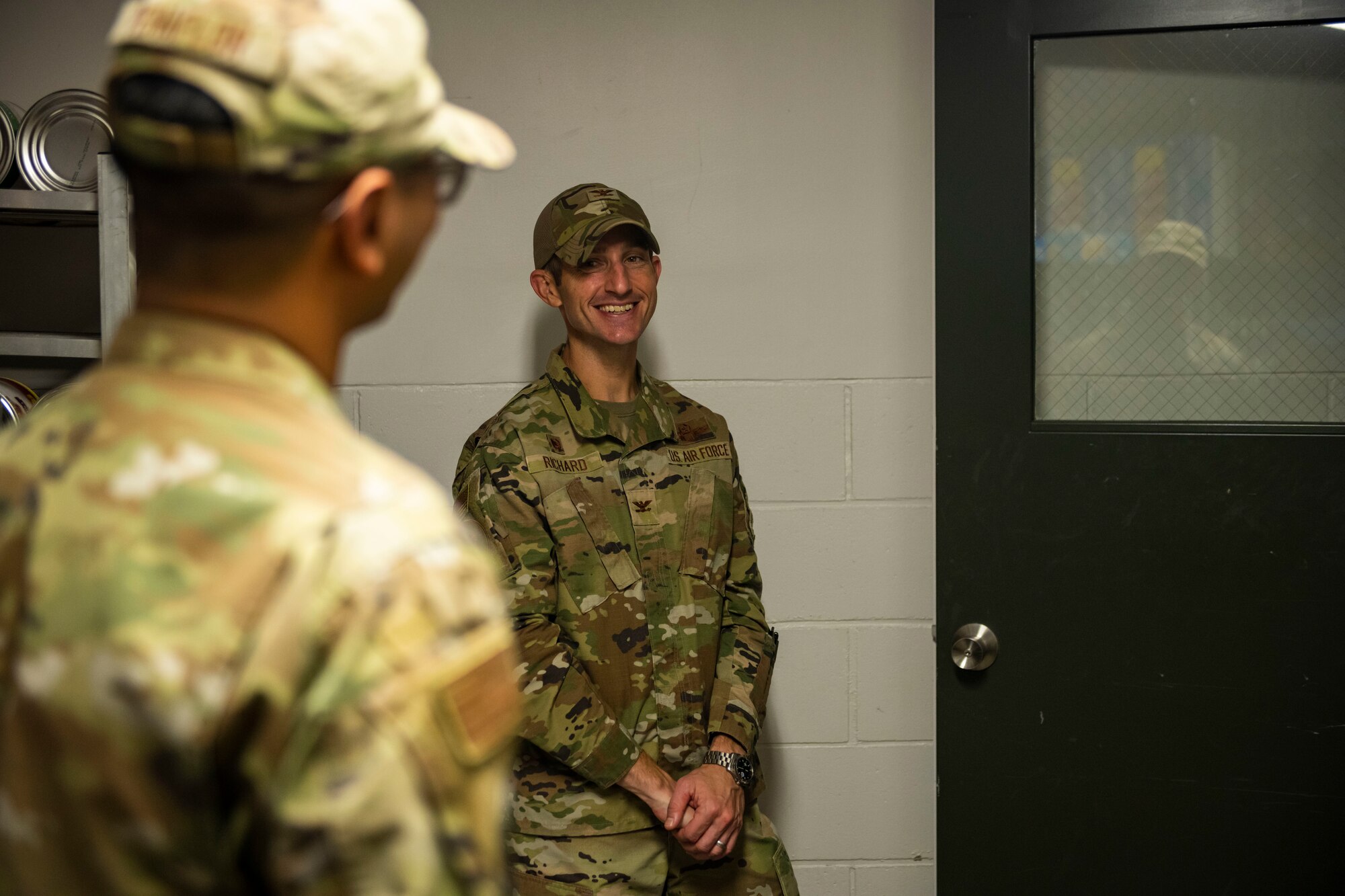 Military members in uniform stand in a dining facility talking.