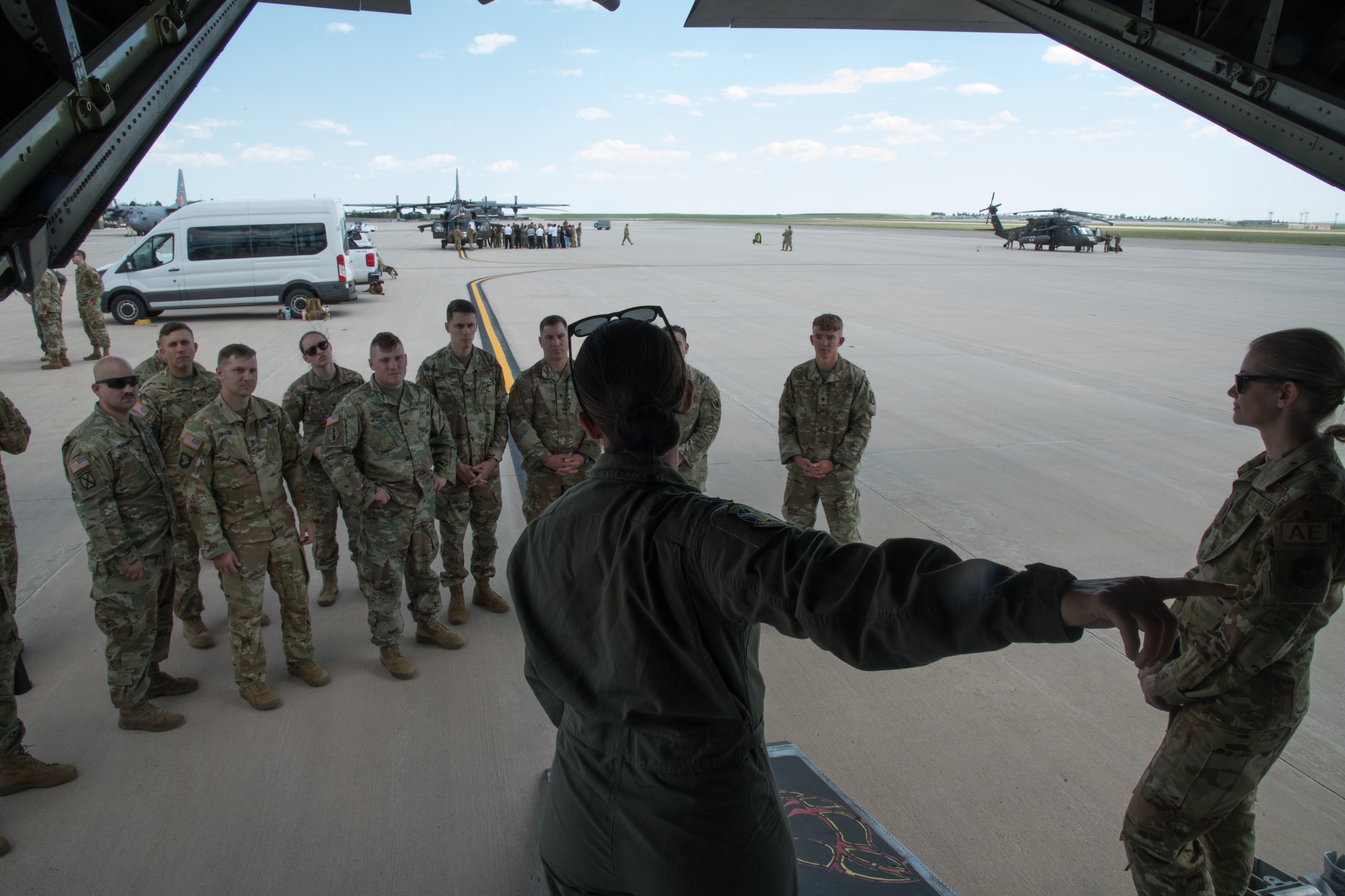 A servicemember in a flight suit points behind her inside of a C-130 aircraft on the flight line while soldiers listen to her. Airmen and soldiers stand near two helicopters in the background.
