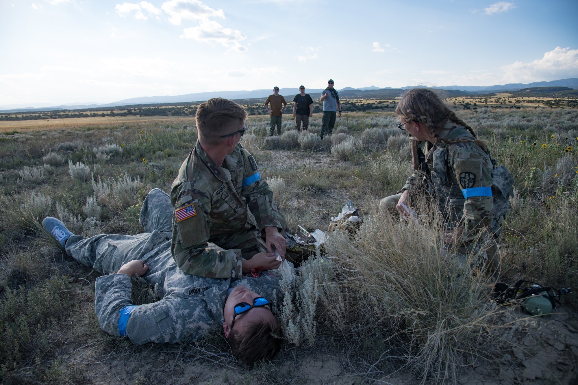 Two uniformed military personnel kneeling in a field, one with his forearm against the chest of a man laying on the ground with fake injuries, looking at three people watching in the background.