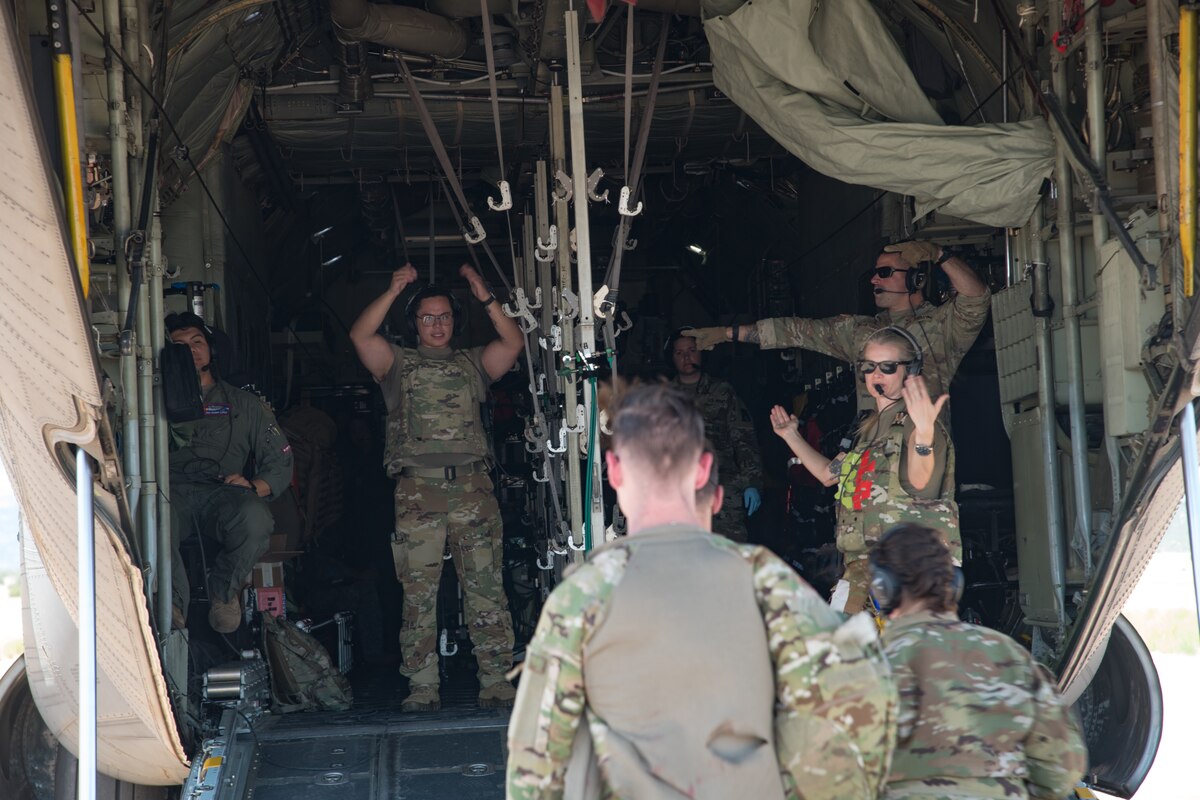 Three uniformed military personnel wave and point to direct a team into the cargo bay of a C-130 aircraft.
