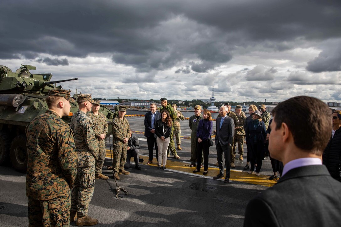 Uniformed service members and several people wearing business attire stand across from each other on the flight deck of a ship.