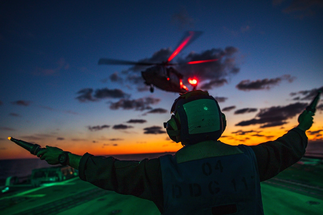 A service member guides a helicopter to land on the flight deck of a ship using traffic wands and hand signals.