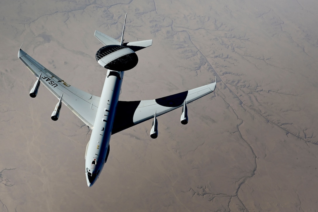 A large aircraft flies above a flat landscape.