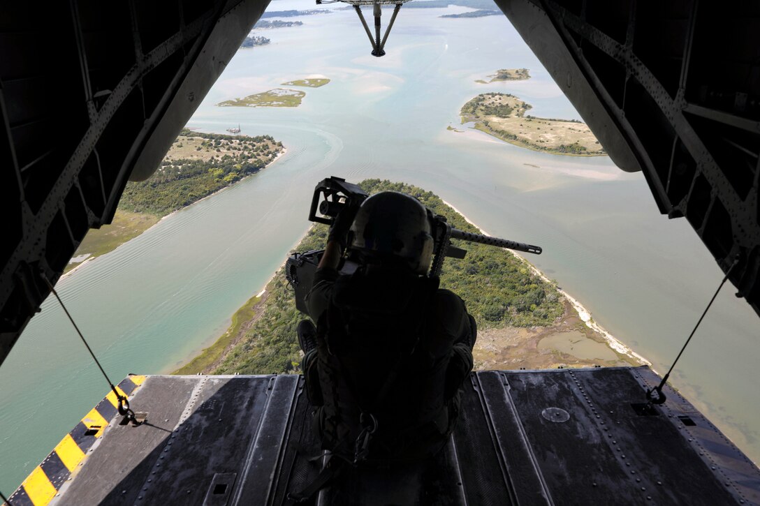A Marine sits behind a mounted weapon in the open doorway of an airborne helicopter.