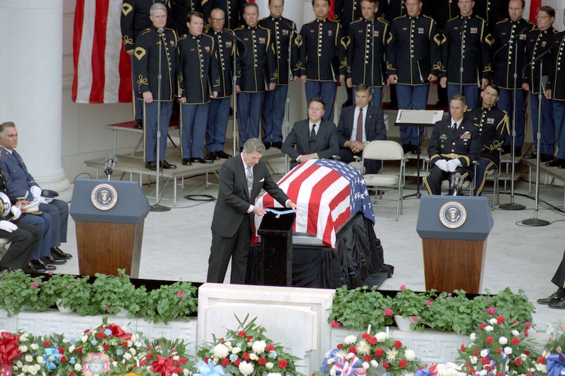 A man places a medal in front of a flag-draped casket. Others stand and sit in the background.