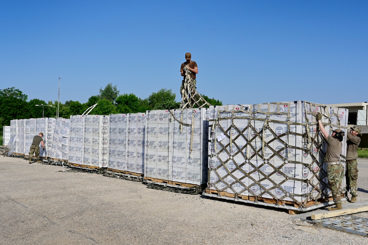 A service member stands atop a pallet contain boxes as others work to unpack other pallets outside.
