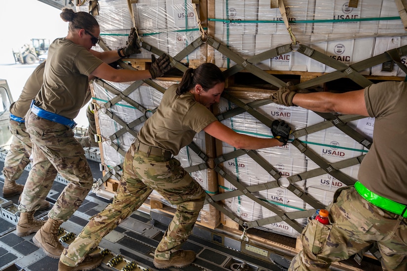 Service members push a pallet of boxes onto an aircraft.
