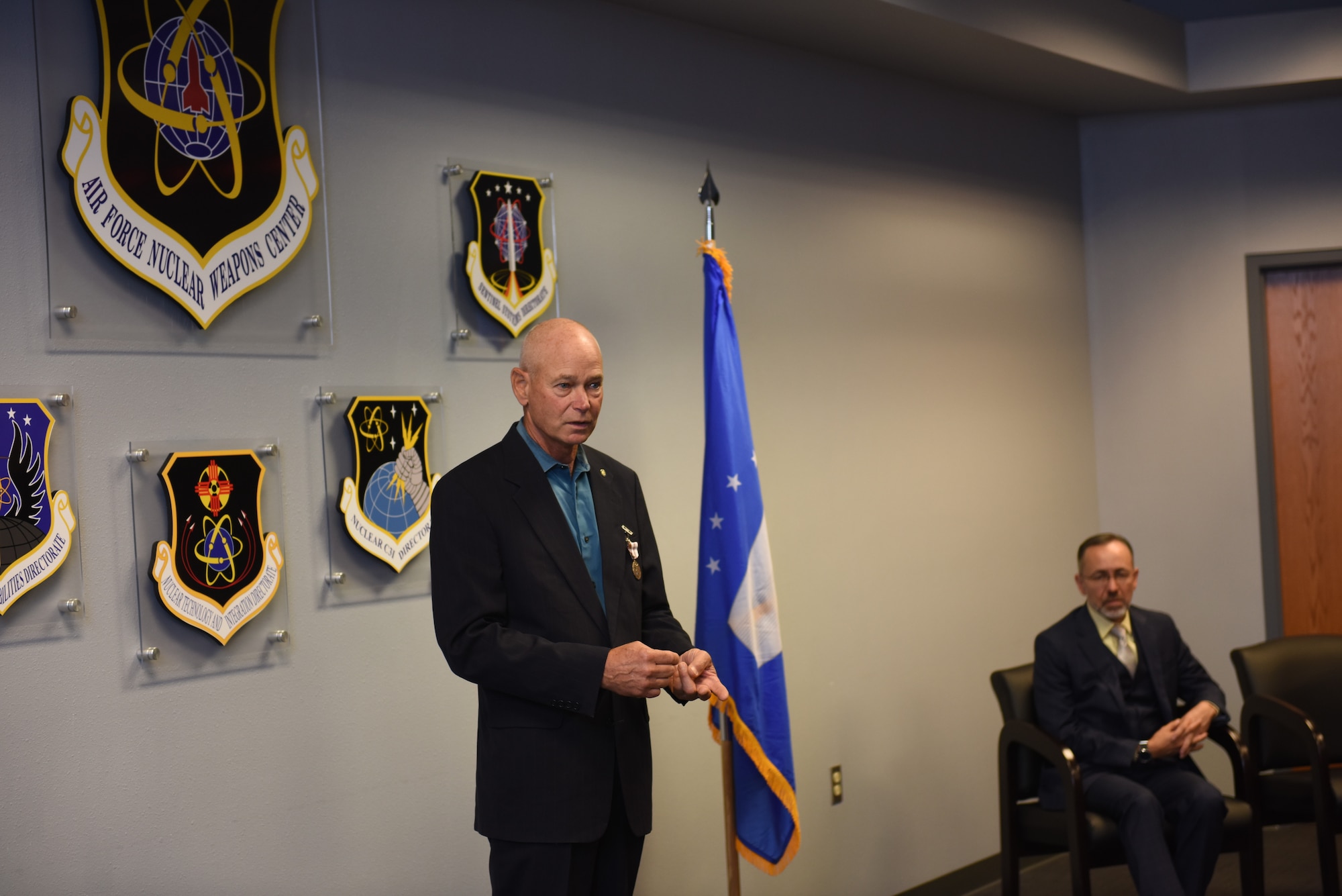 A man in a suit stands in front of a blue flag and a wall of government crests