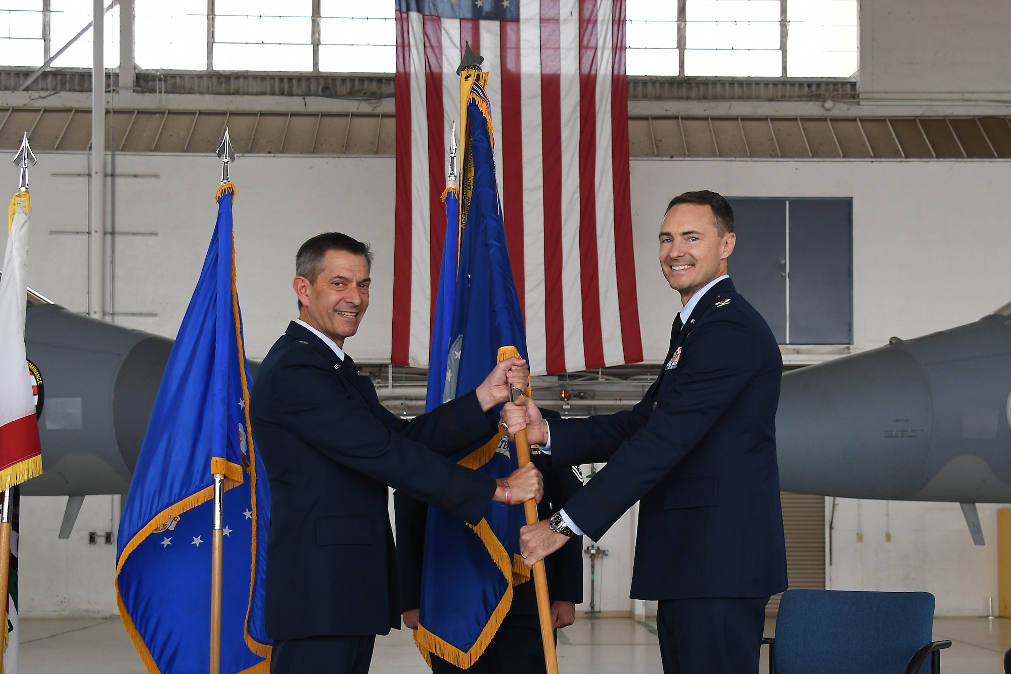 Two men stand wearing the Air Force service dress uniform (blue suit) facing the photo frame holding an blue Air Force guidon (flag) between them