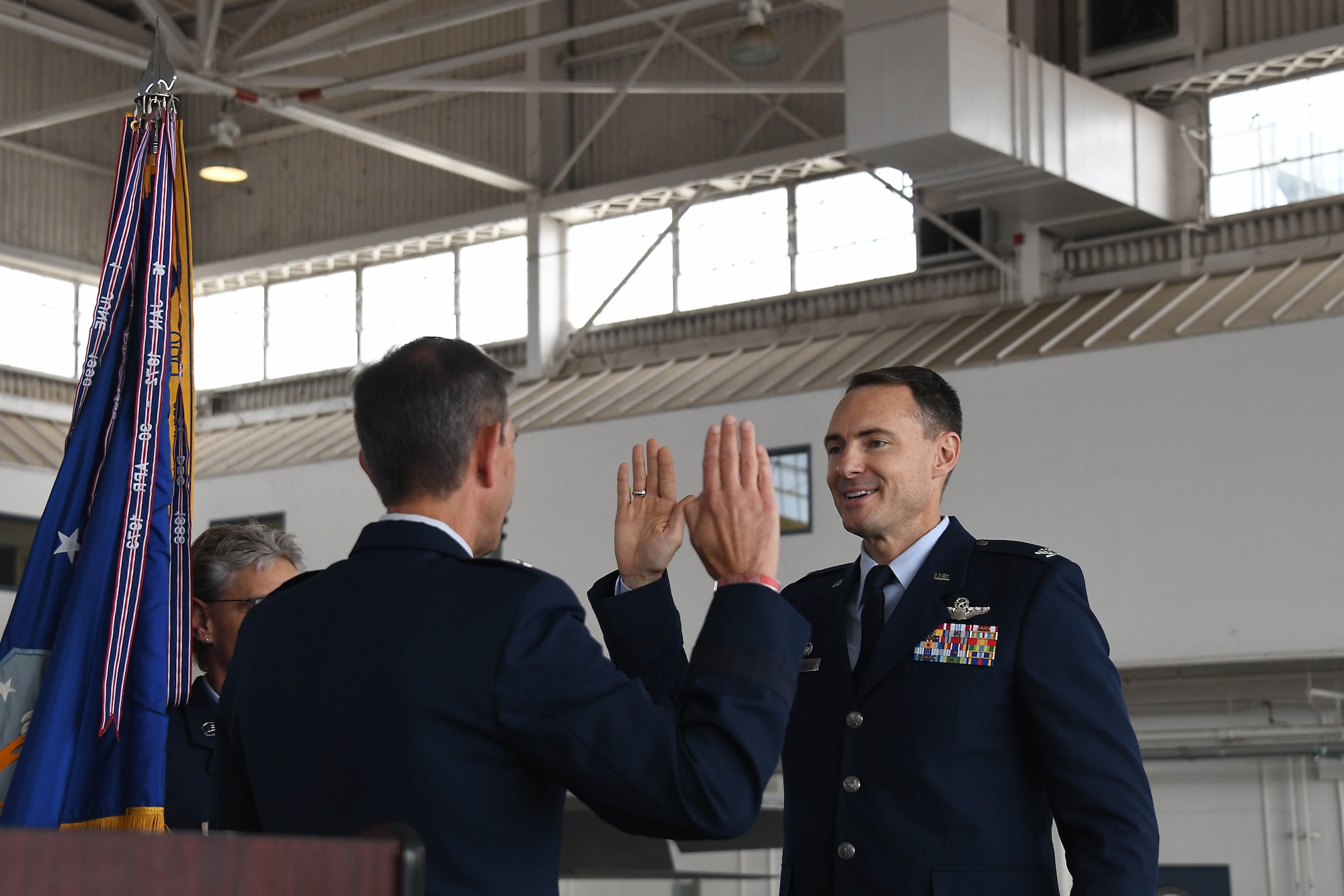From right, a man in an Air Force service dress uniform (blue suit) holds up his right hand to swear in to a position. On the left, a man with in a similar blue suit stands with his back to the front of the photo frame.