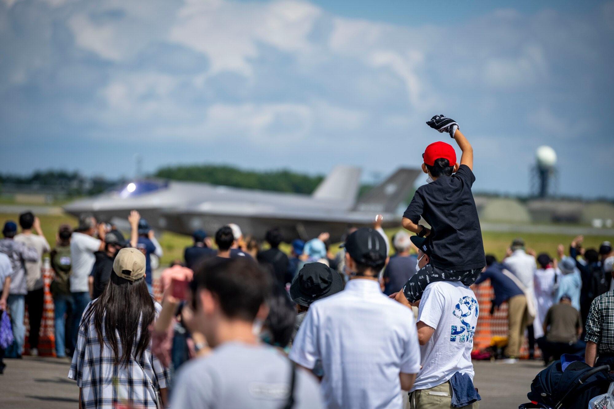 A Japanese child sits on fathers shoulders as they wave to a military aircraft passing by.