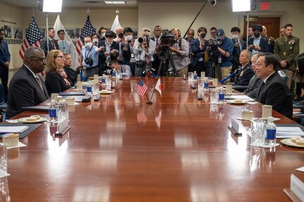 People sit at a conference table as journalists gather at the end with cameras and notepads.