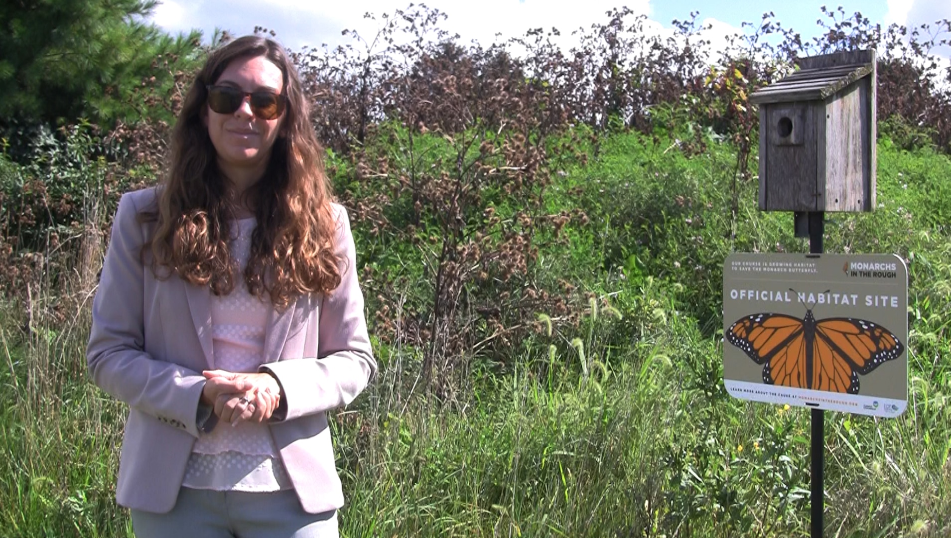 A woman in a pink shirt and grey suit with long brown hair stands in front of a Monarchs in the Rough sign in front of a prairie area on the DSCC golf course.