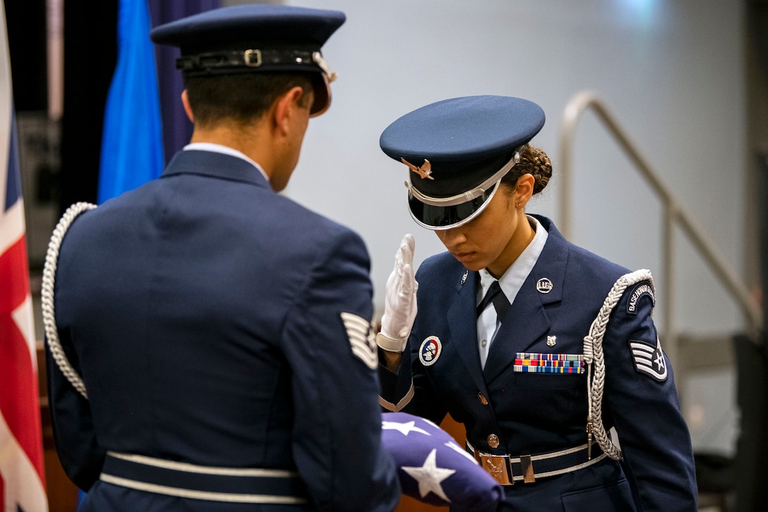 An airman salutes a folded flag being held by another airman.