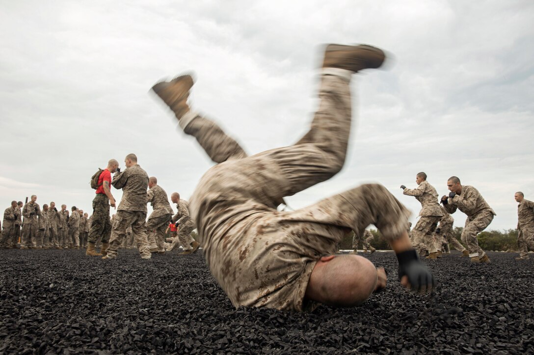 A Marine Corps recruit rolls on the ground while fellow recruits conduct martial arts training in the background.