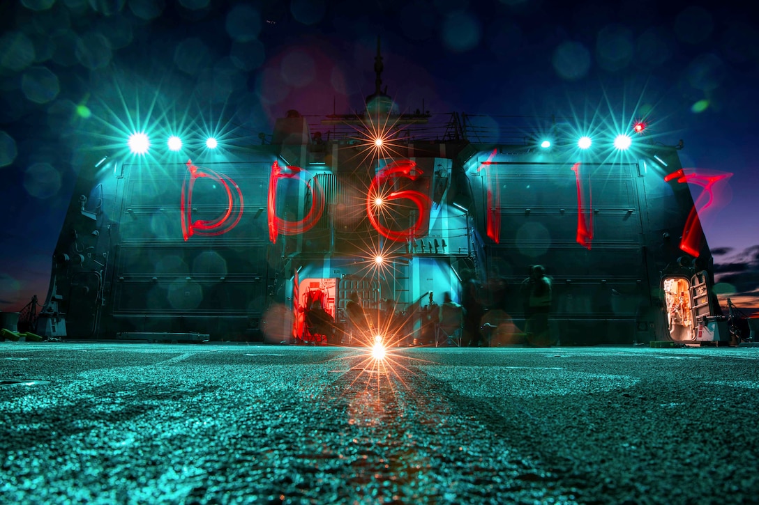 Sailors sit on the flight deck of a ship illuminated by headlights with red writing reflected on a screen.