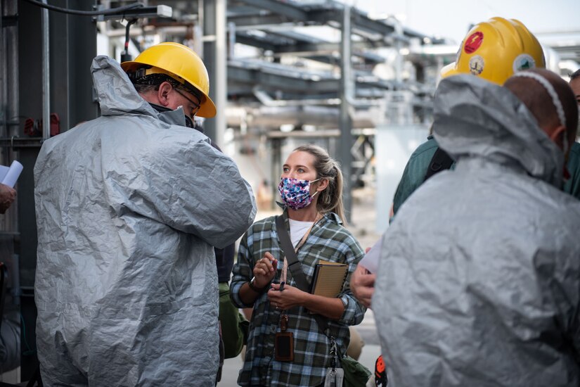 A woman with a notepad and a facemask talks to a group of workers wearing personally protective equipment.
