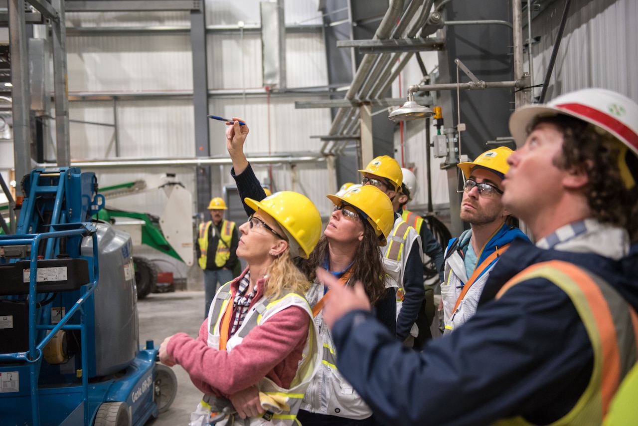 A woman wearing a hardhat gestures toward something in a warehouse while a group of workers also wearing hard hats look on.