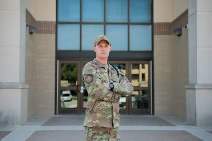 Man stands in front of a building while posing with his arms across his chest. He is looking at the camera.