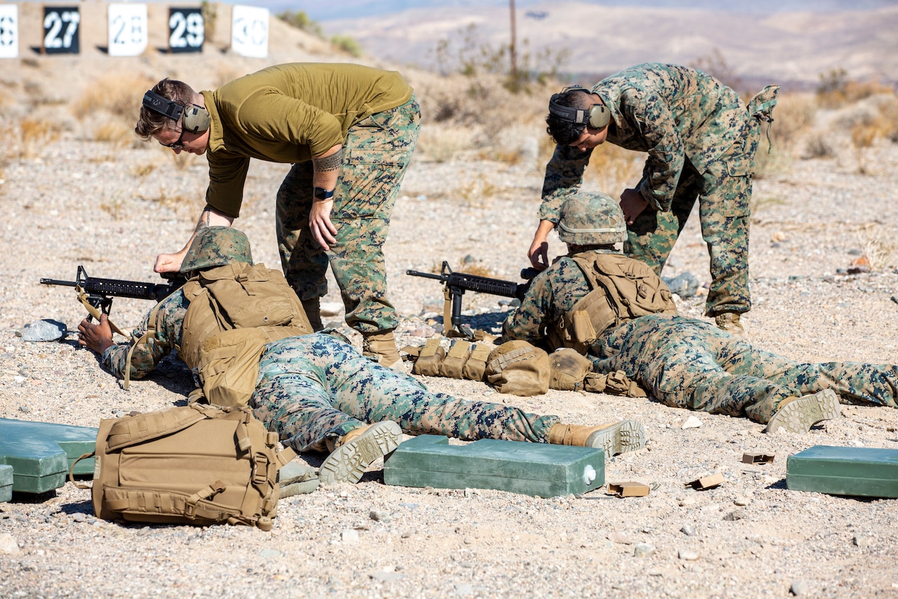Two Marines hold weapons as they lie in the dirt on their stomachs; two other Marines bend down and touch the weapons.