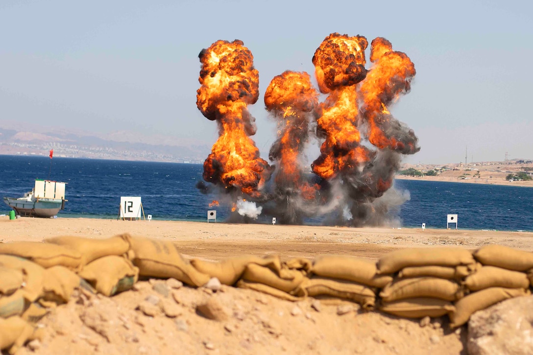 Smoke and flames rise from an explosion on a beach next to a small boat.