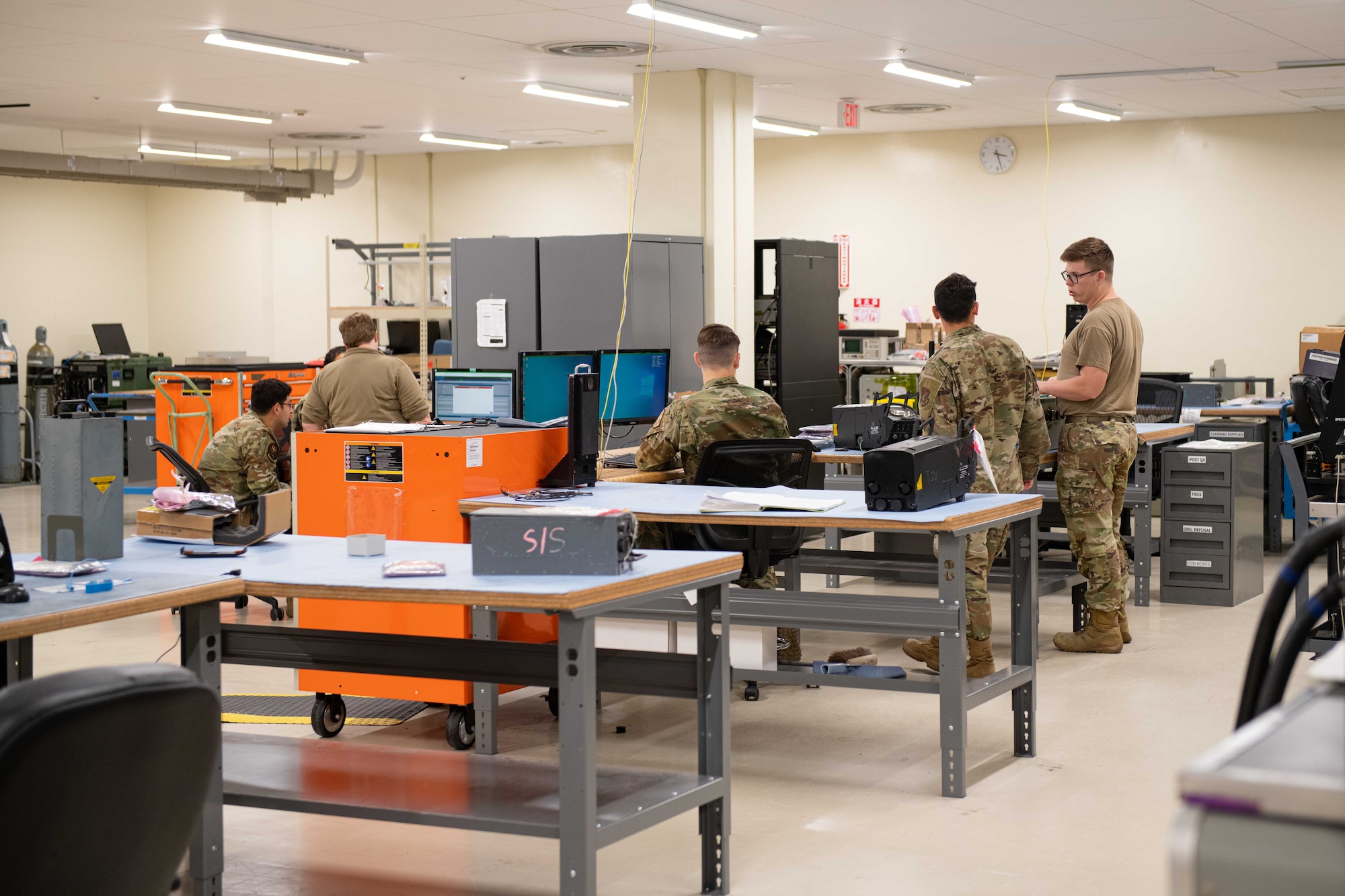 A group of Airmen work around desks and tables in the avionics section of a shop
