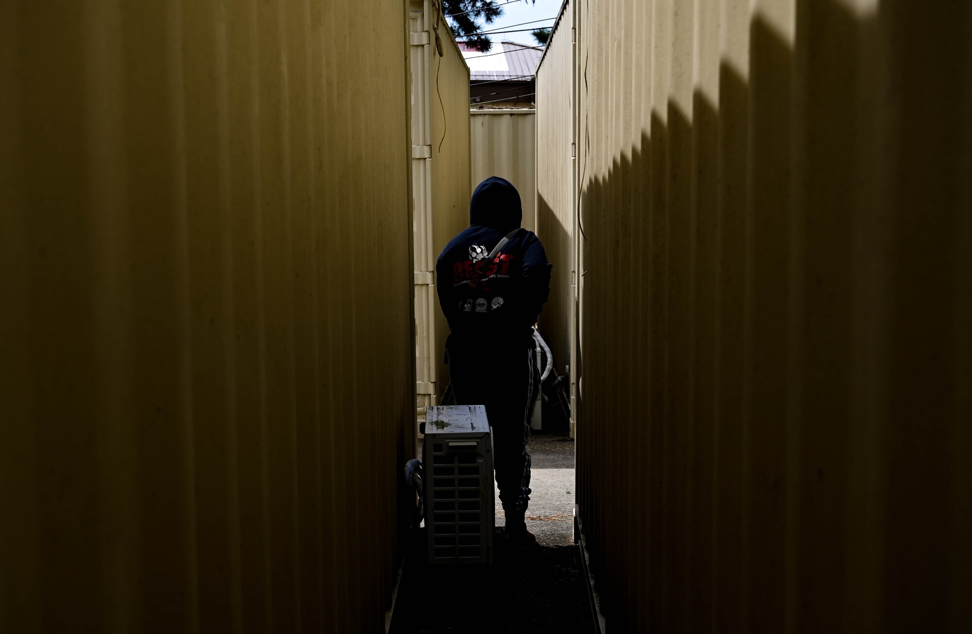 Airman dressed as civilian walks between cargo boxes with weapon.
