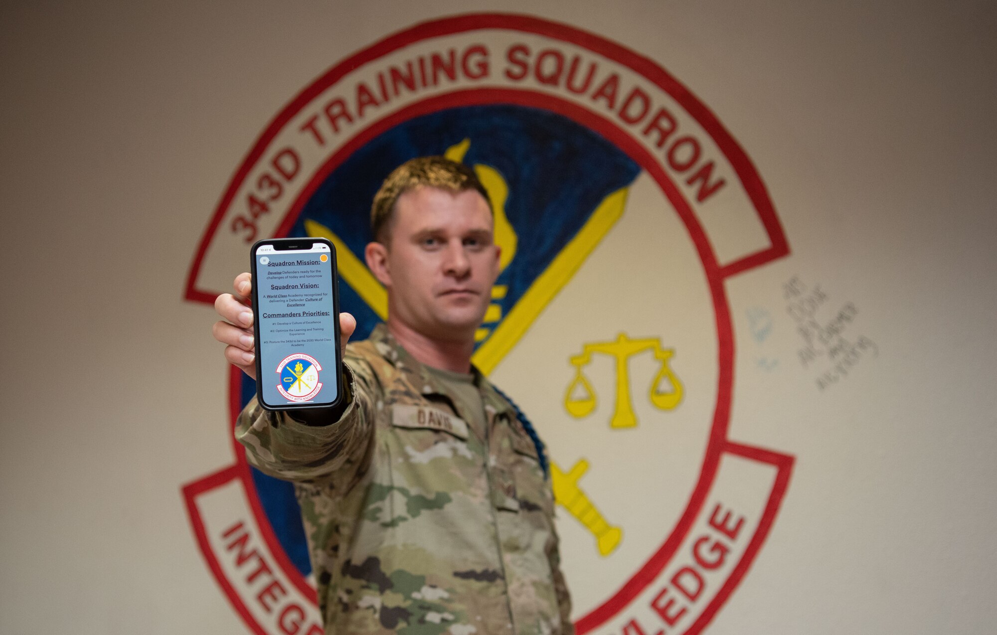 Man stands in front of squadron logo painted on the wall. He is holding up a phone, which is what the camera is focused on, while a webpage is populated on his phone.