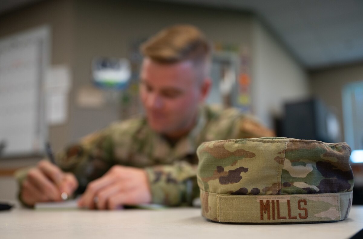A hat with the name "Mills" sewed on sits in front of an Airman writing notes