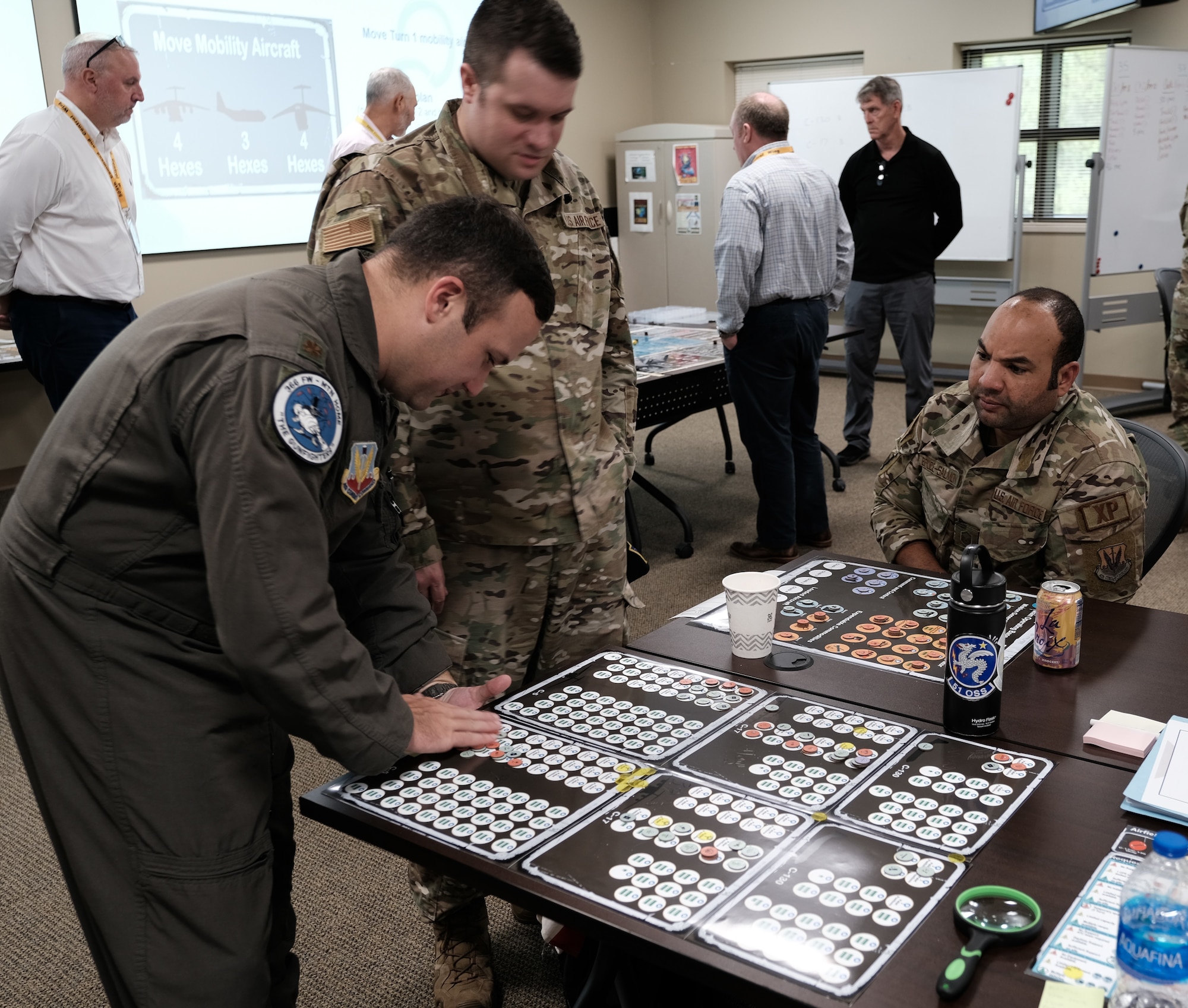 photo of US Military members training using a board game with USG civilians in the background standing around a table
