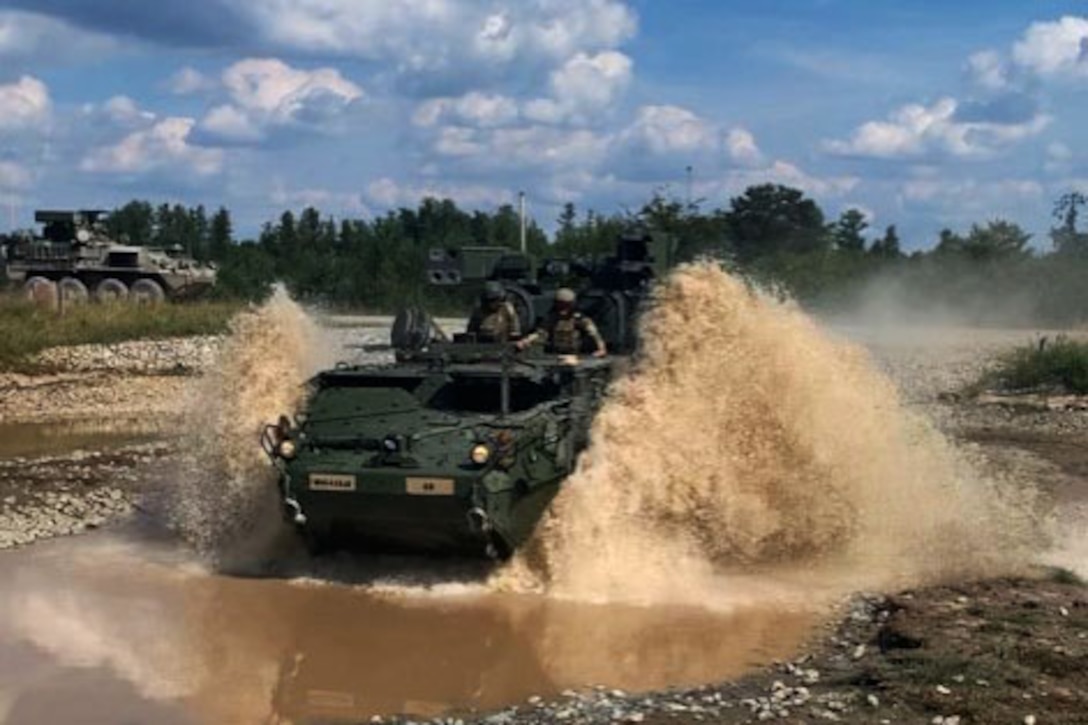 Two soldiers ride a tank through a mud puddle.