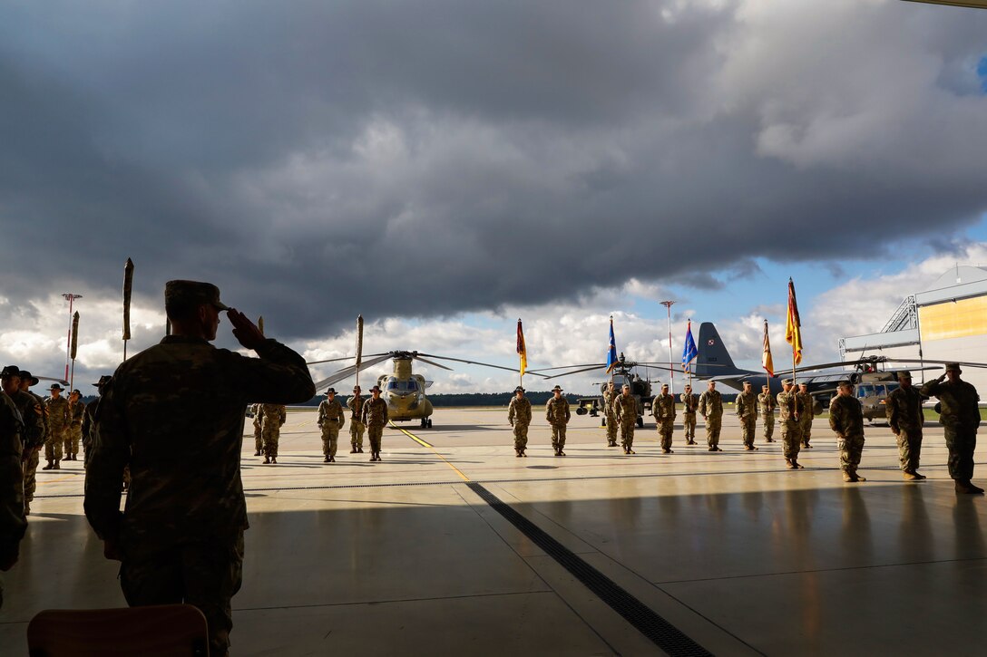 A uniformed service member salutes a formation of soldiers outside with military aircraft in the background.