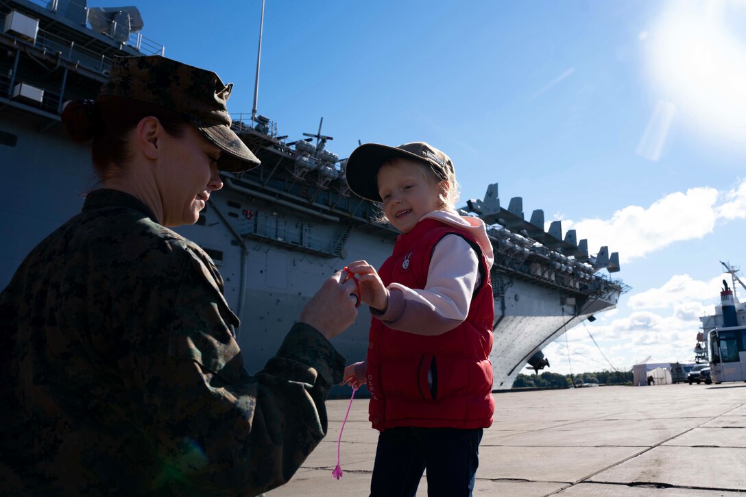 A  kneeling uniformed service member plays with a young girl on a pier with a ship in the background.