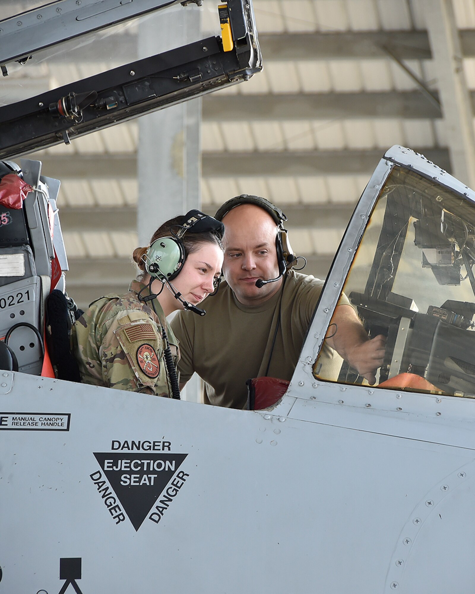 Female Airman sits in cockpit of A-10, looking at controls while male Airman stands over her shoulder pointing to controls he is familiarizing her with.
