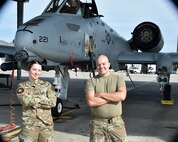 Two Airmen, female left and male on right posing with arms crossed in front of chests, looking at camera, A-10 Thunderbolt II aircraft parked behind them in background.