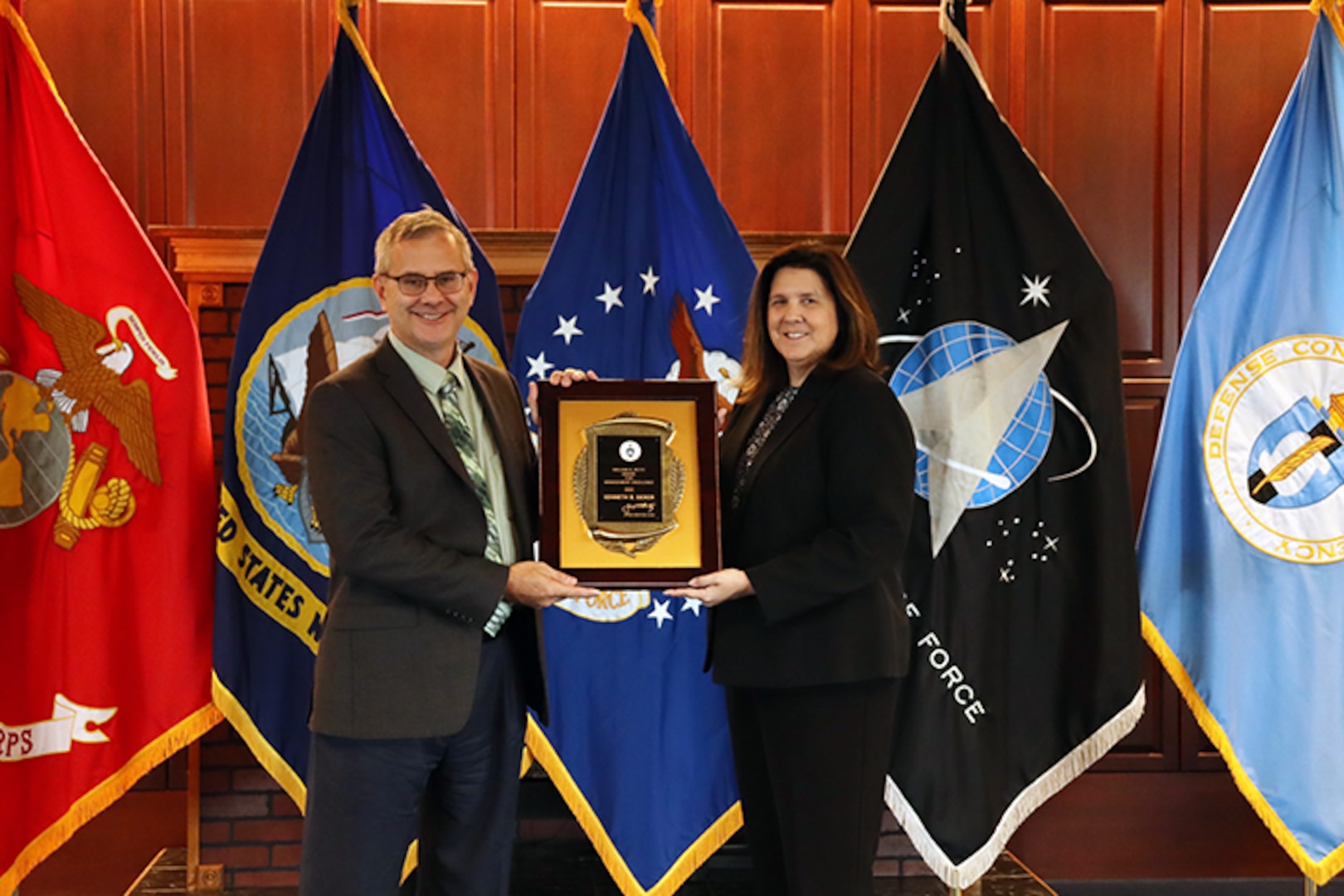 man and woman holding award with service flags in background