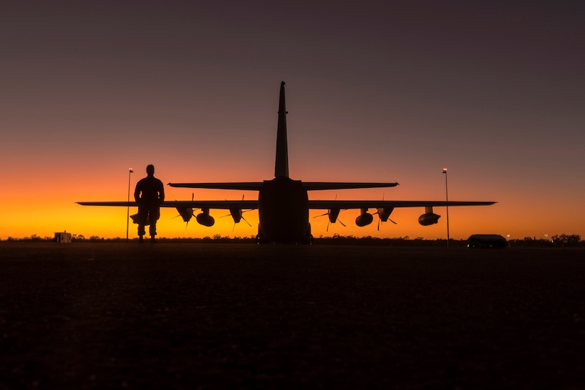 A service member stands near an aircraft under a sunlit sky as seen in silhouette.