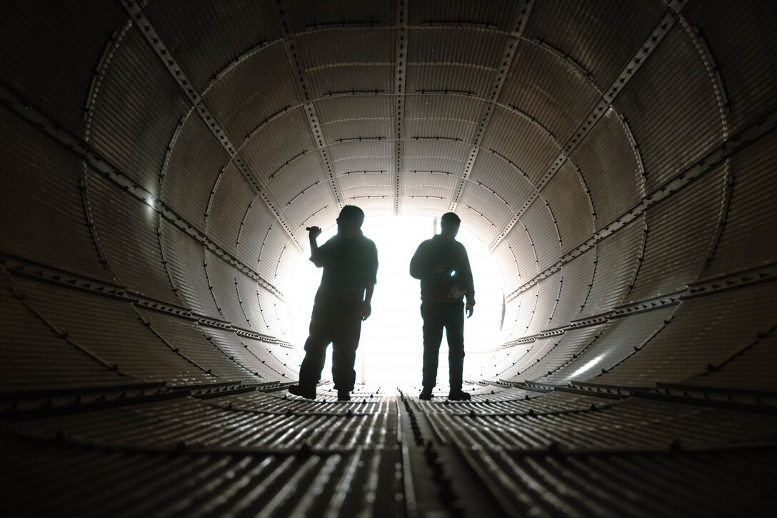 Two men using flashlights walk in a large metal tube.